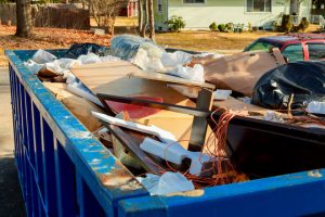 A blue dumpster filled with various household debris including cardboard boxes, plastic bags, and other discarded materials in what appears to be a residential area. 