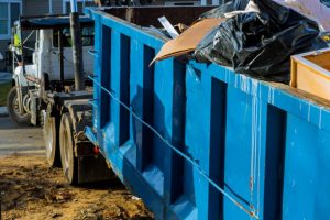A close-up view of a large blue industrial dumpster partially filled with debris, showing its ribbed metal construction and part of the truck chassis it sits on.
