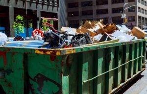 A large green industrial dumpster filled with mixed debris including cardboard boxes and other materials sits on a city street with office buildings visible in the background.