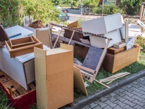 A pile of discarded furniture and household items, including wooden cabinets and shelves, stacked on a grassy area near a sidewalk