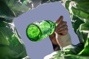 a person disposing of a green glass bottle, viewed from inside a green plastic bag against a clear sky.