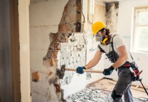 A construction worker wearing protective gear (including a yellow hard hat, face mask, and ear protection) is using a sledgehammer to demolish a brick wall during what appears to be a home renovation project.