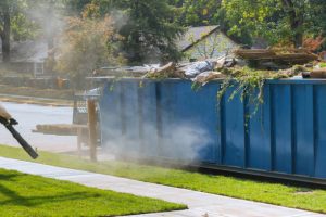 A blue dumpster sits in a residential area with steam or smoke visibly rising around it on a sunny day with autumn trees in the background.
