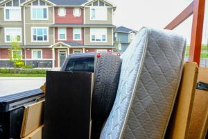 A mattress, tire, and other discarded items are piled up in front of modern townhouses or condominiums, appearing to be either moving day debris or bulk waste waiting for disposal.