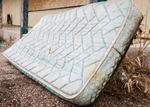 An old, weathered mattress with a quilted light blue pattern lies discarded on a gravel surface next to some dried vegetation.