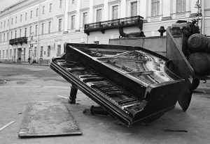 A black and white photograph shows a damaged grand piano lying on its side on a street in front of a classical building with ornate architecture.
