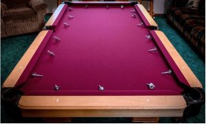 A pool table with burgundy felt and wooden trim is shown with screws or bolts laid out around its perimeter, suggesting it's in the process of being assembled or repaired.