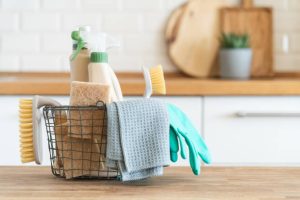 A wire basket filled with cleaning supplies including rubber gloves, spray bottles, scrub brushes, and a dishcloth sits on a wooden counter in a kitchen setting.