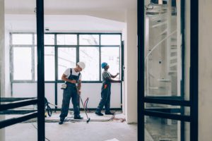 Two construction workers in overalls are working in an unfinished room with large windows, with one examining the floor and another inspecting the window area.