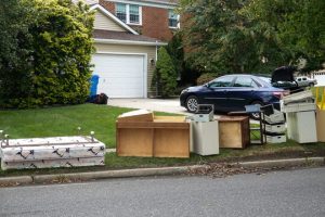 This image shows various discarded furniture and cardboard boxes placed at the curb for pickup in front of a suburban home with a white garage door and a dark-colored car in the driveway.
