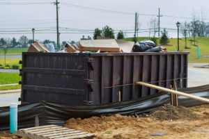 A large brown metal dumpster filled with construction debris sits on a dirt area with black plastic sheeting at its base, with utility poles and green fields visible in the background.