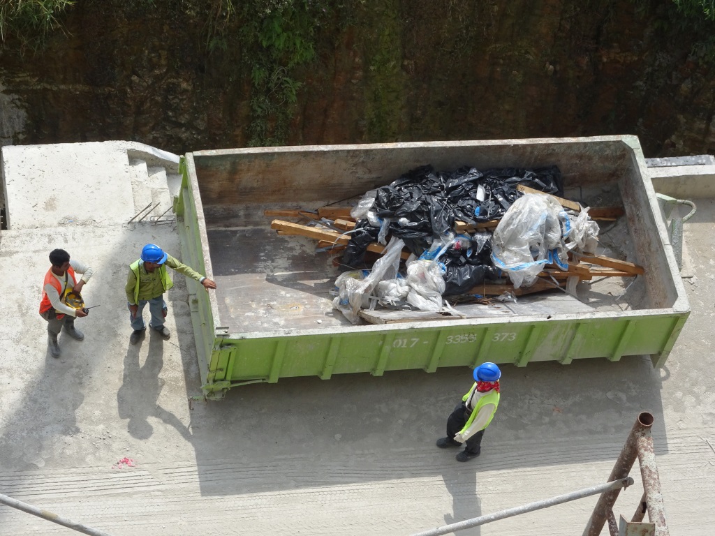 Construction workers in safety gear are standing around a large green dumpster filled with debris, trash bags, and wooden materials at a job site.RetryClaude can make mistakes. Please double-check responses.